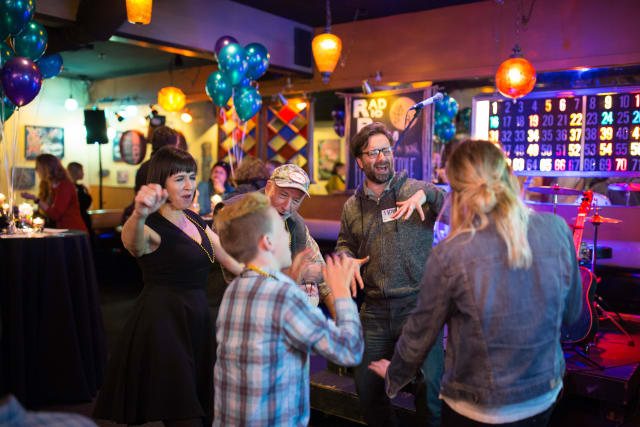 Group of diverse attendees enjoying the Love Bomb 2017 event, dancing and celebrating in a lively venue decorated with balloons and colorful lights, supporting My Voice Music nonprofit.